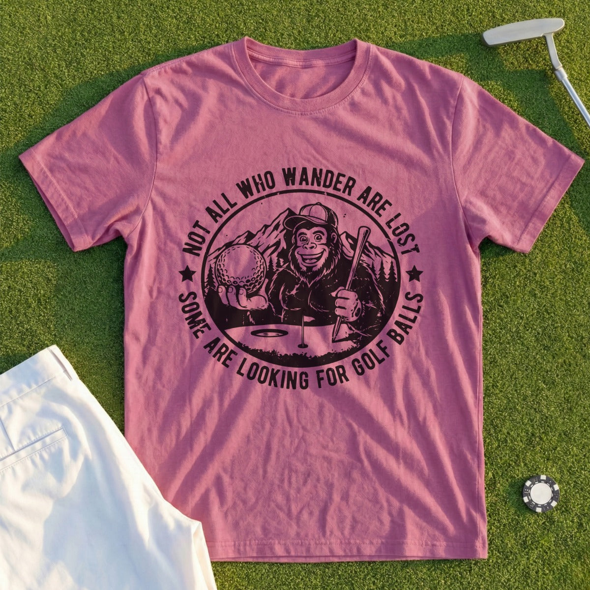 Pink t-shirt with Bigfoot holding a golf ball and club, text "Not all who wander are lost, some are looking for golf balls."