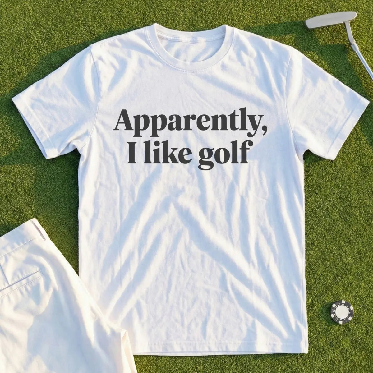 White t-shirt with "Apparently, I like golf" text, laid on grass with golf club and ball.