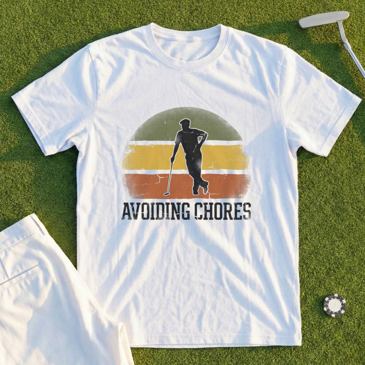 White t-shirt with "AVOIDING CHORES" and golfer graphic, laid on grass.