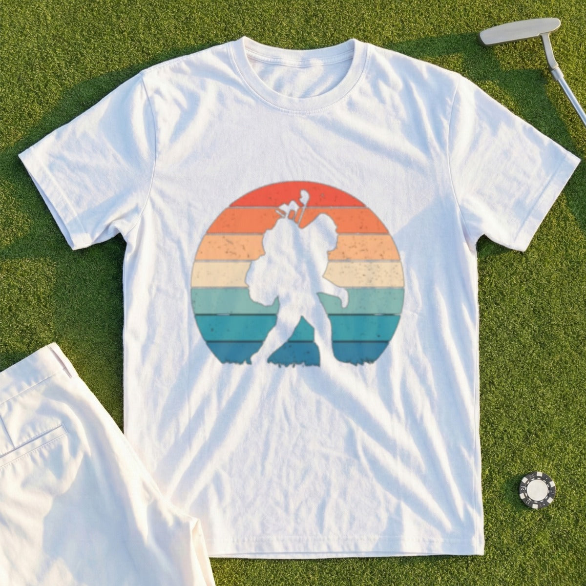 Bigfoot carrying golf clubs against a retro sunset graphic on a white t-shirt.
