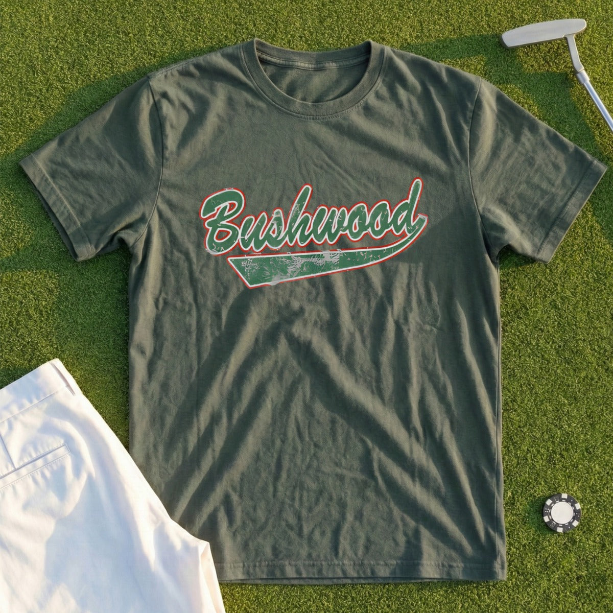 Green t-shirt with "Bushwood" in red and white cursive text, lying on grass.