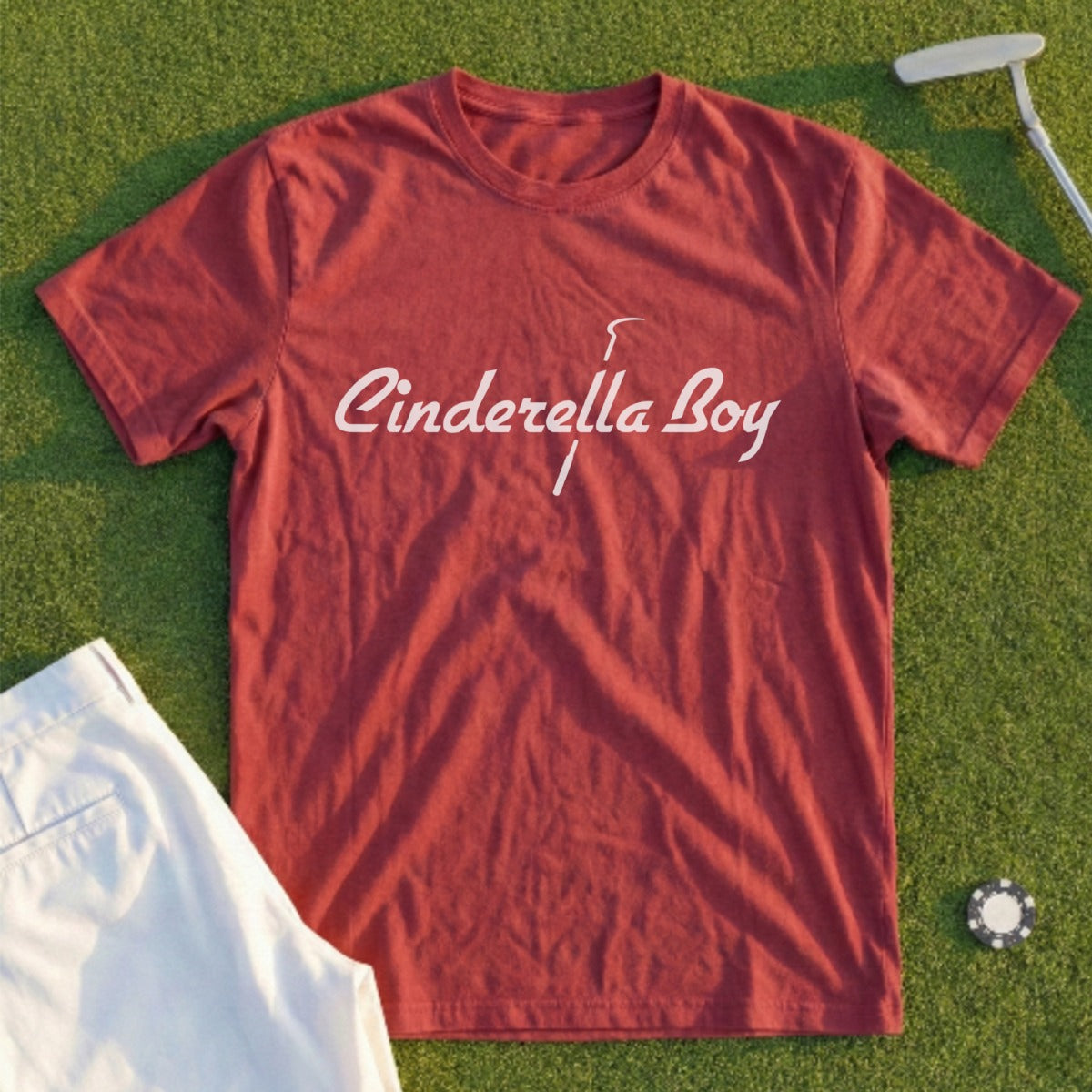 Red t-shirt with "Cinderella Boy" text, laid on grass with golf putter.