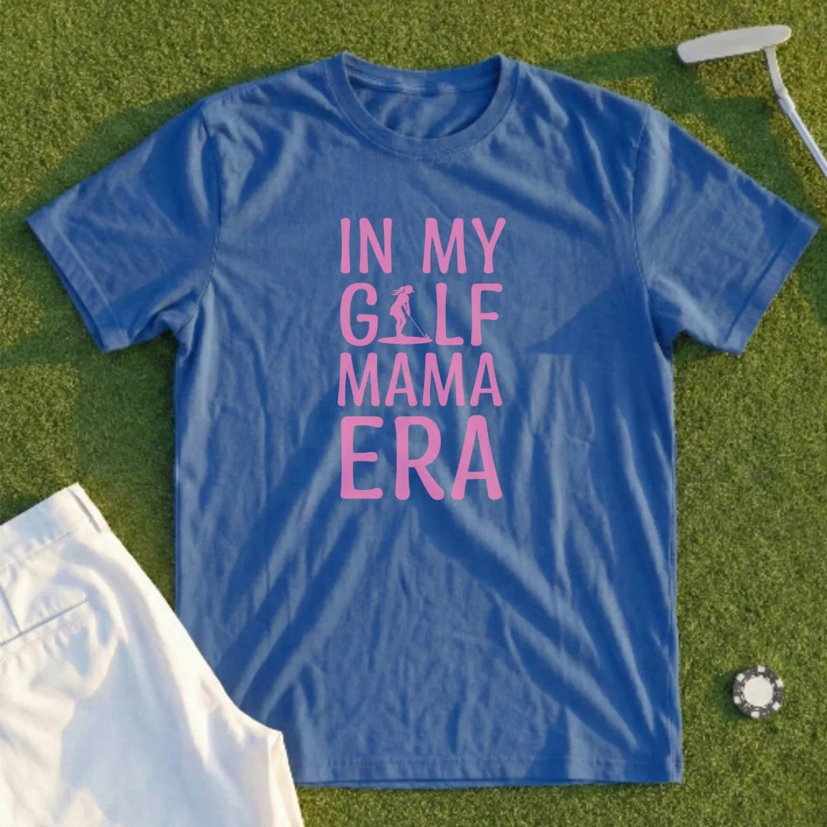 Blue t-shirt with "In My Golf Mama Era" text, laid on grass near golf accessories.