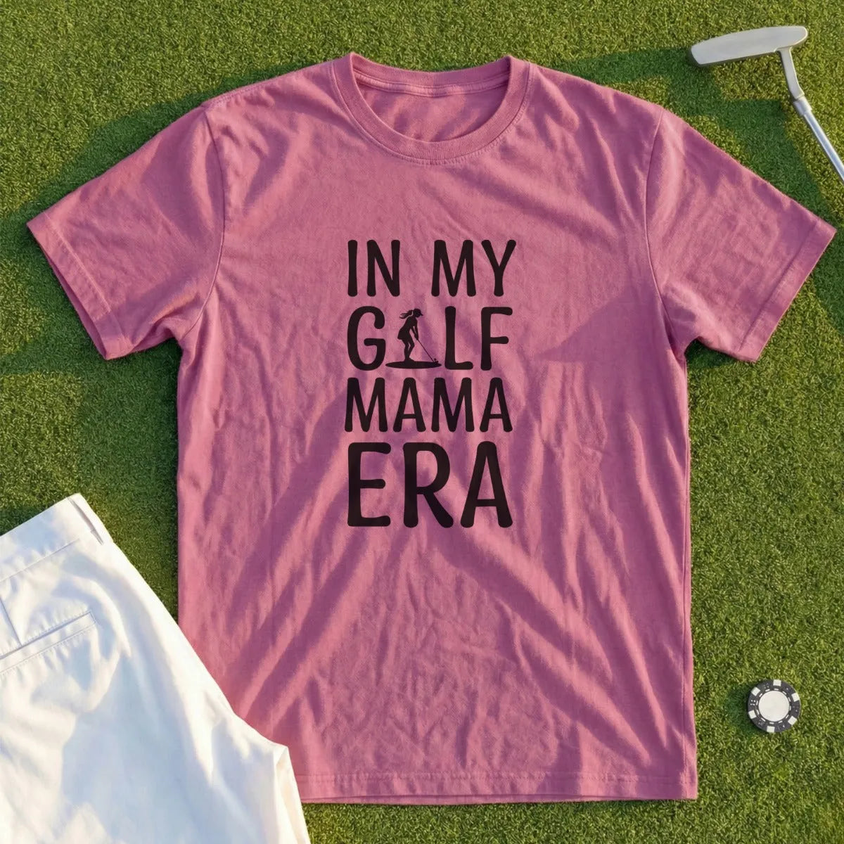 Pink t-shirt with "In My Golf Mama Era" text, laid on grass with golf club and shorts.