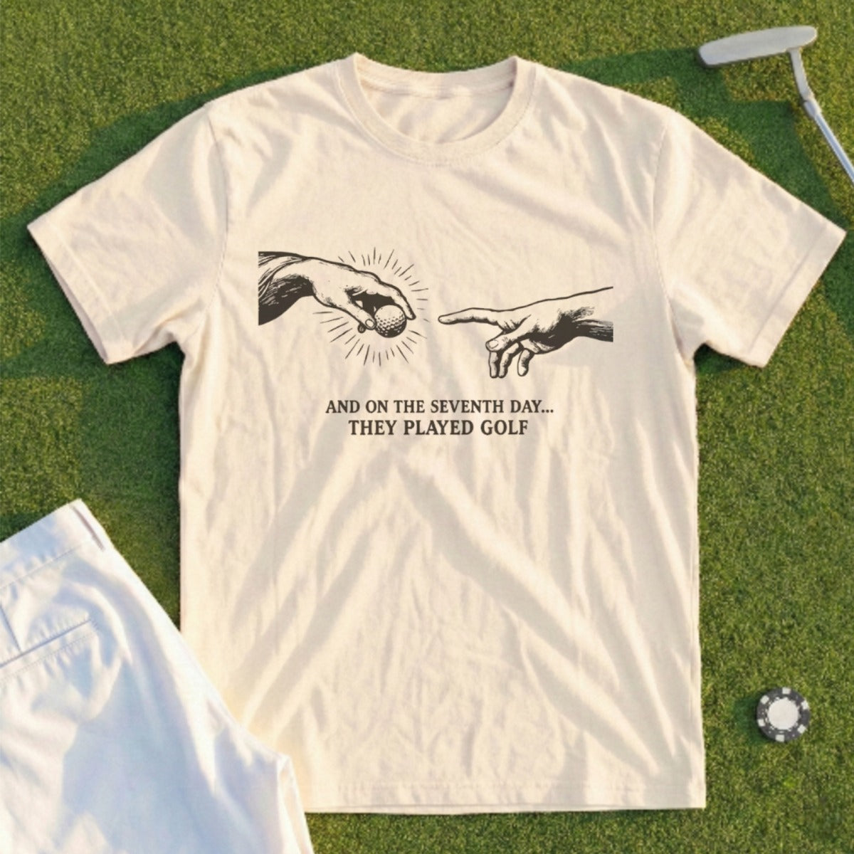 Cream t-shirt with "The Creation of Adam" hands reaching for golf ball, text reads "AND ON THE SEVENTH DAY... THEY PLAYED GOLF".