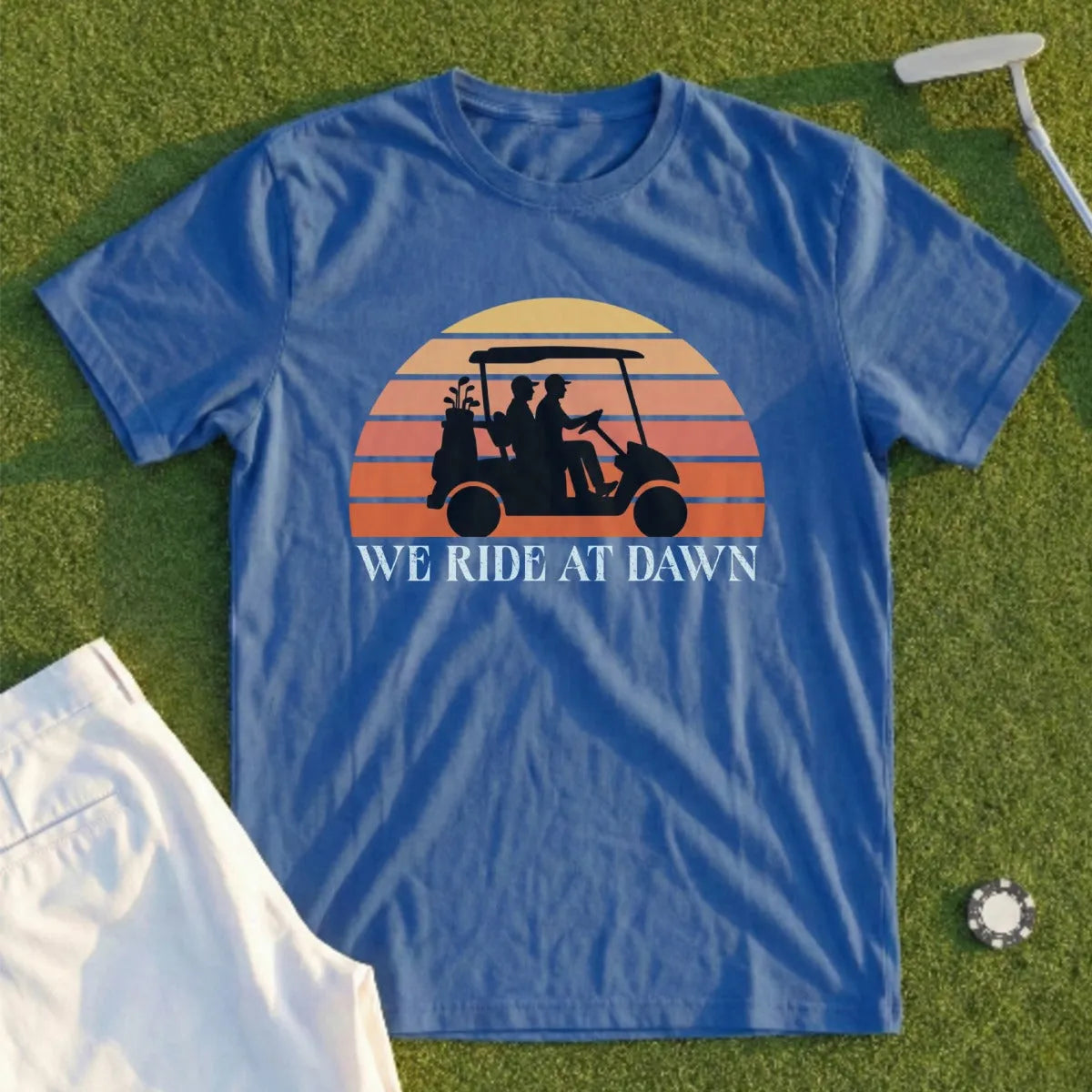 Blue t-shirt with a sunset graphic of golfers in a cart and "WE RIDE AT DAWN" text.