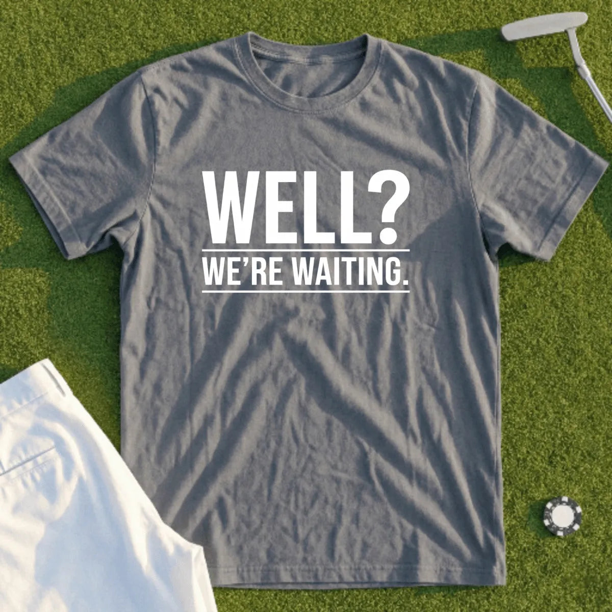 Gray t-shirt with "WELL? WE'RE WAITING." text, on grass with golf accessories.