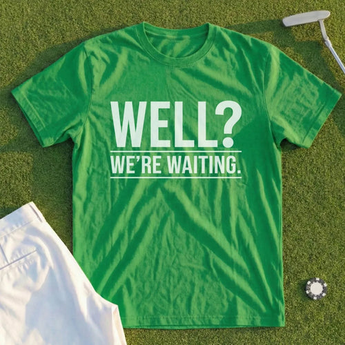 Green t-shirt with white text "WELL? WE'RE WAITING." on a golf course.