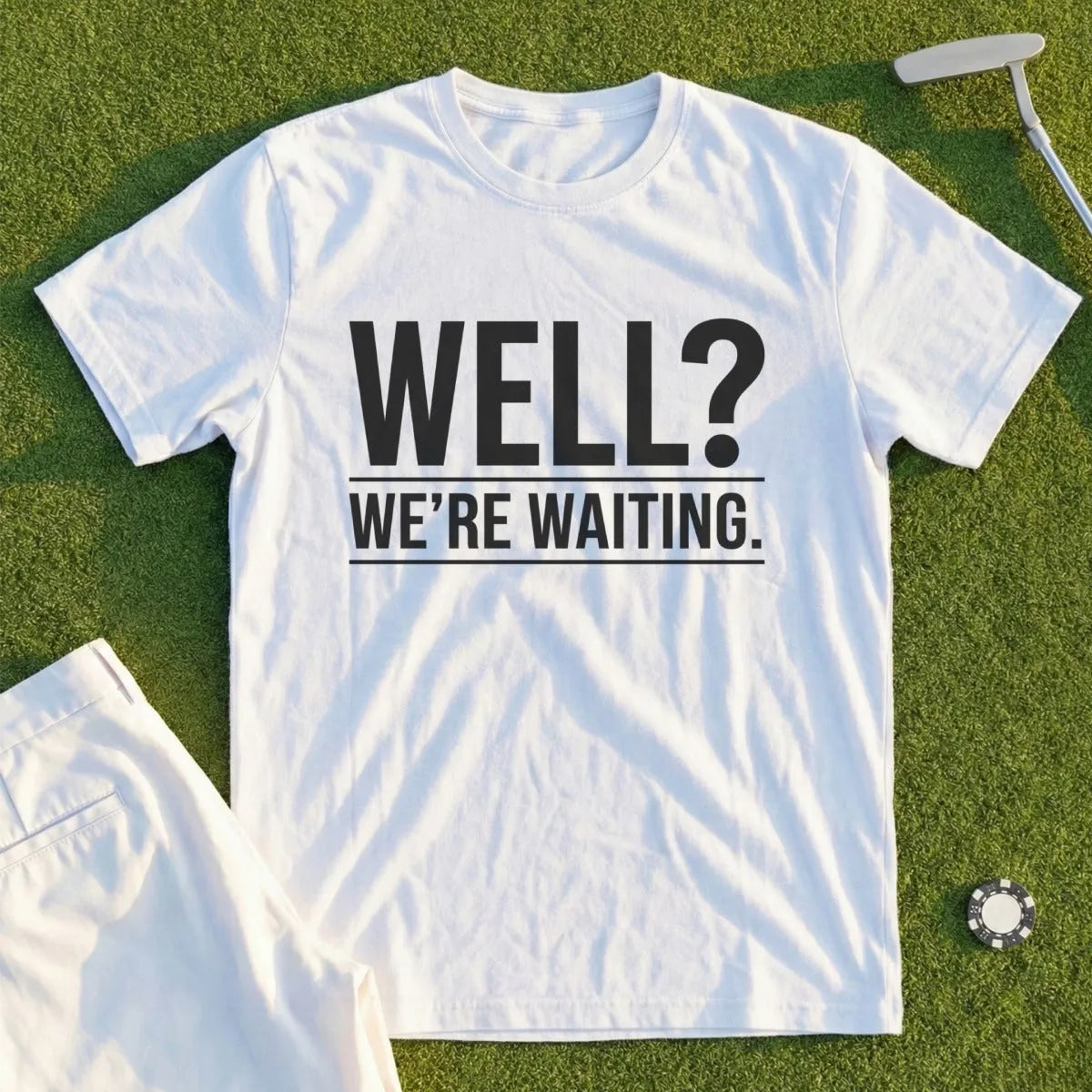 White t-shirt with black text "WELL? WE'RE WAITING." on a grassy surface.